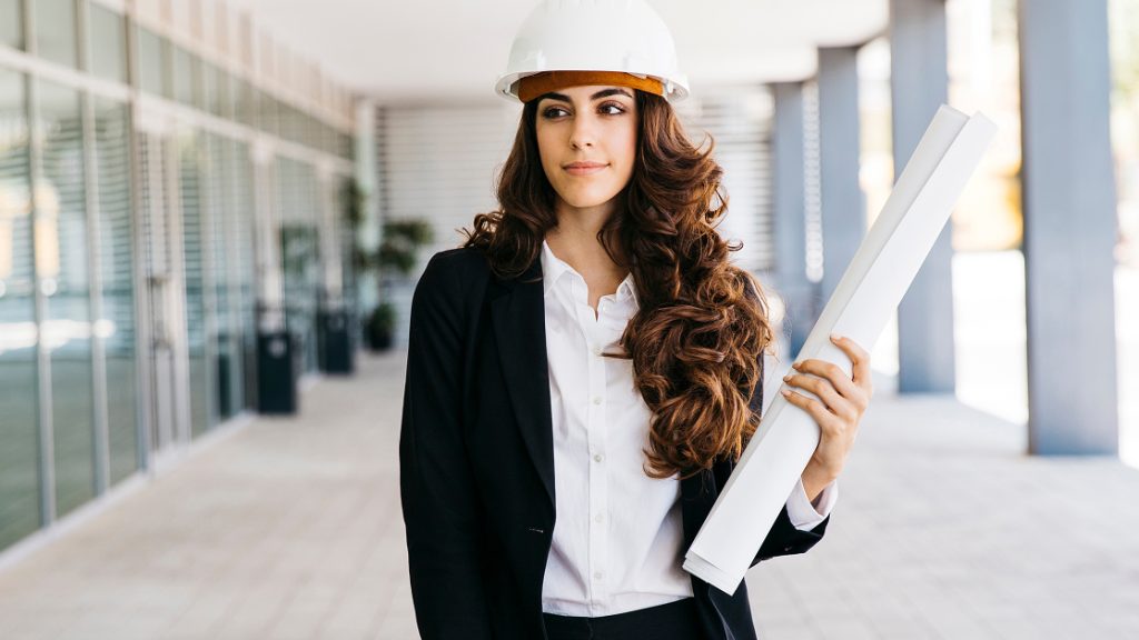 A woman wearing a white hard hat and a business suit holding rolled-up blueprints with a confident expression, embodying the drive of AEC leaders and the Nemetschek Academic Mission supported by Graphisoft.